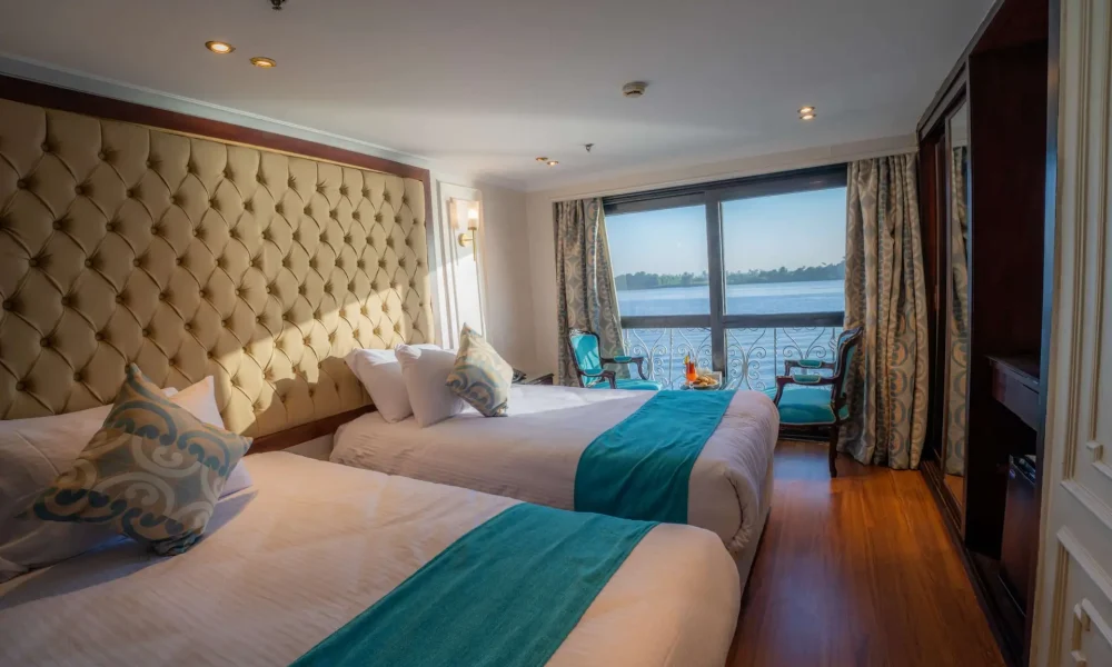 Spacious cruise ship cabin with twin beds, plush headboard, large window, and turquoise accents overlooking the river