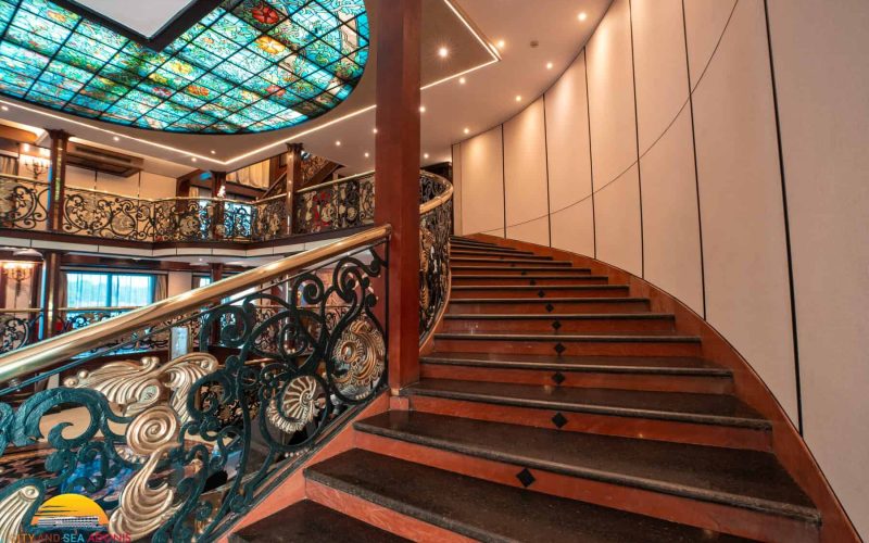 Grand staircase aboard the M/S City & Sea Adonis with ornate railings and stained glass ceiling.