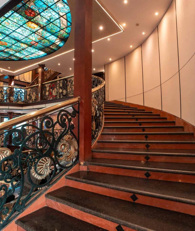 Grand staircase aboard the M/S City & Sea Adonis with ornate railings and stained glass ceiling.