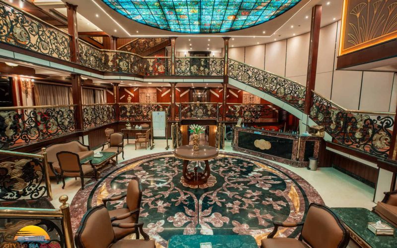 Luxurious two-level atrium lounge aboard the M/S City & Sea Adonis with ornate railings and turquoise skylight.
