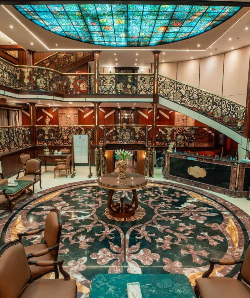 Luxurious two-level atrium lounge aboard the M/S City & Sea Adonis with ornate railings and turquoise skylight.