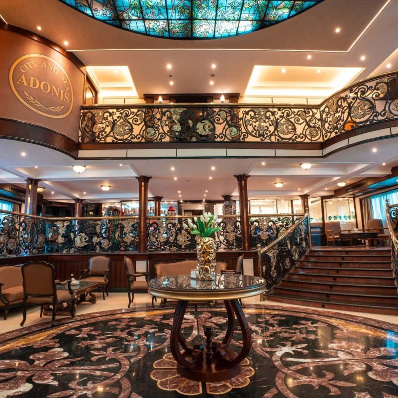 Elegant atrium lobby aboard the M/S City & Sea Adonis with ornate railings and central staircase.