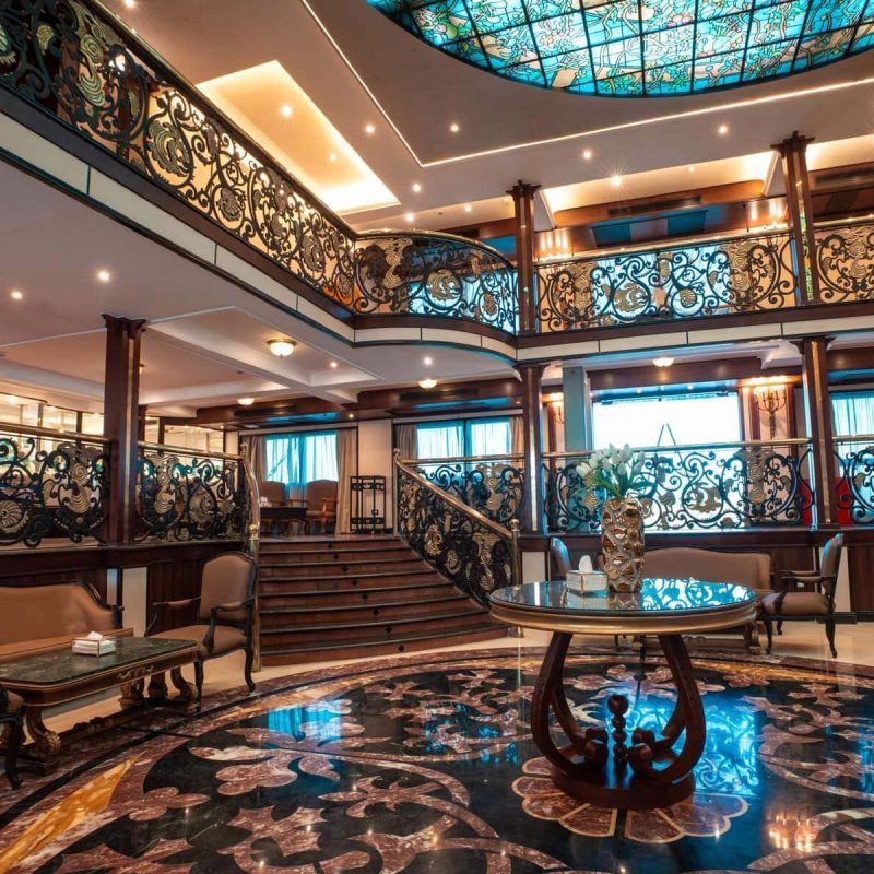 Elegant multi-level atrium lounge aboard the M/S City & Sea Adonis with ornate railings and skylight.