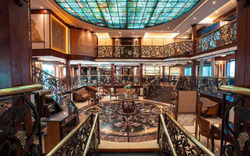 Elegant multi-level atrium lounge aboard the M/S City & Sea Adonis with ornate railings and turquoise stained glass ceiling.