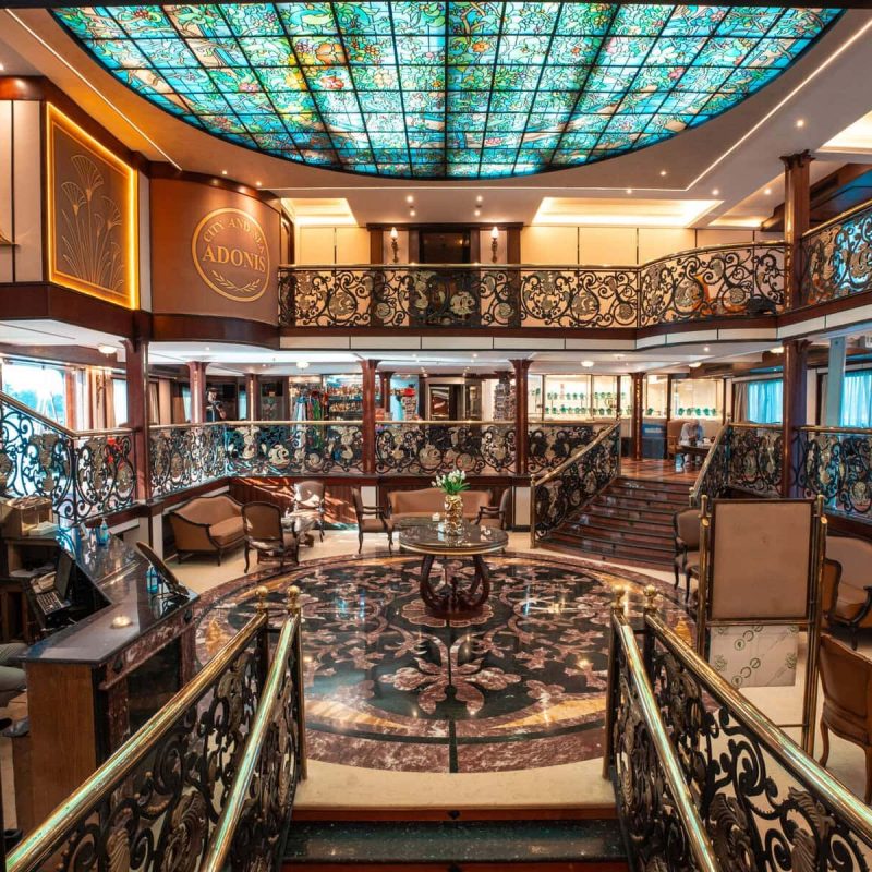 Elegant multi-level atrium lounge aboard the M/S City & Sea Adonis with ornate railings and turquoise stained glass ceiling.