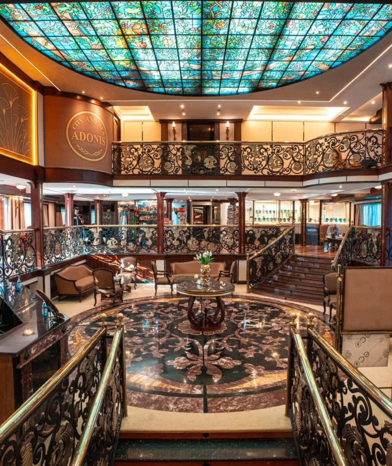 Elegant multi-level atrium lounge aboard the M/S City & Sea Adonis with ornate railings and turquoise stained glass ceiling.