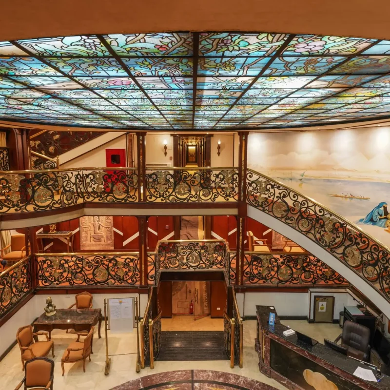 Grand cruise ship reception area with a stained glass ceiling, ornate iron railings, and classical Egyptian murals