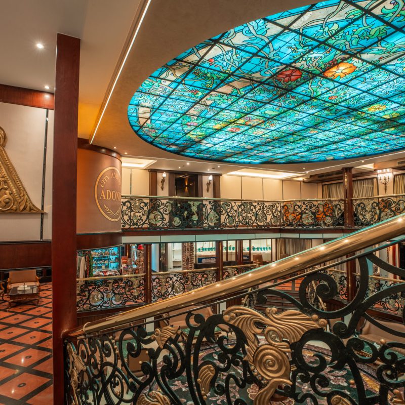 Grand staircase aboard the M/S City and Sea Adonis with ornate railings and turquoise stained glass ceiling.