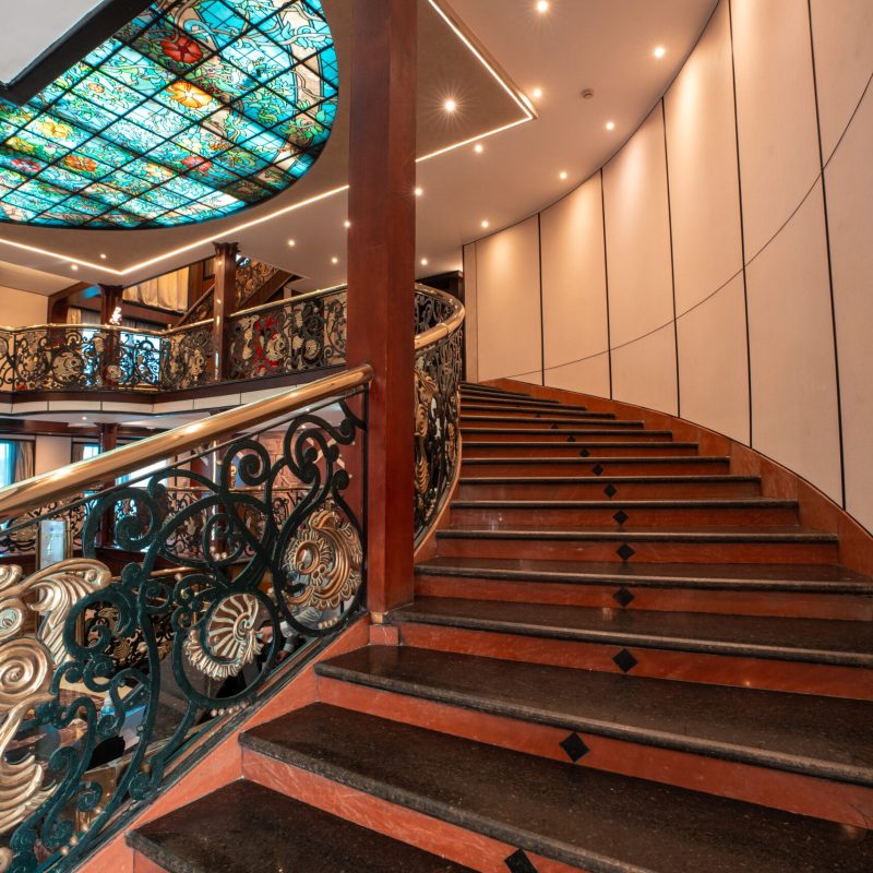 Grand staircase aboard the M/S City and Sea Adonis with ornate railings and stained glass ceiling.