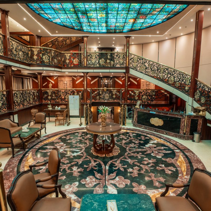 Luxurious two-level atrium lounge aboard the M/S City and Sea Adonis with ornate railings and turquoise skylight.